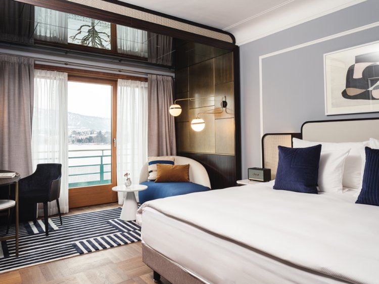 Luxuriöses Schlafzimmer im Art-déco Stil mit grossem, cremefarbigem Bett rechts und einem Fenster mit Aussicht auf den Zürichsee in der Mitte.