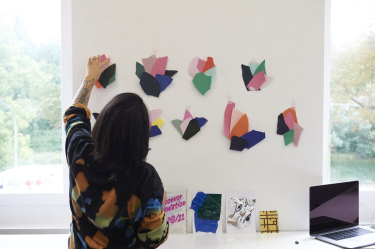 Mara Tschudi vor weisser Wand mit verschiedenen Farb-Arrangements aus bunten Papierfetzen