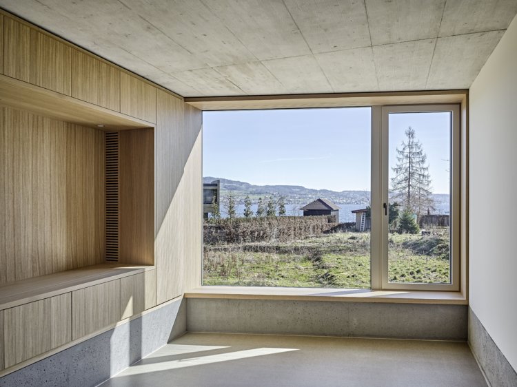 Blick aus Wohnzimmer mit Betondecke und Holzeinbauelementen links durch ein grosses Fenster auf den Zürichsee.