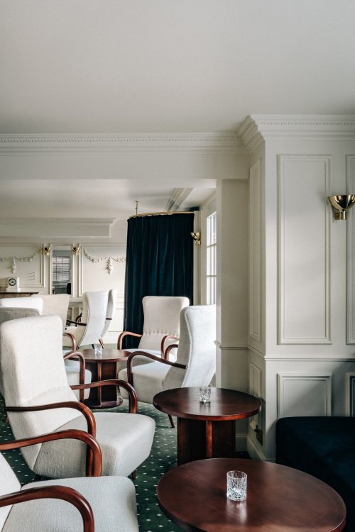 Sessel mit weissen Bouclé-Bezügen und dunklen Holzgestellen auf einem dunkelgrünen Teppich, umgeben von Art-Deco Lampen, schaffen ein gemütliches Ambiente in der Hotelbar des Experimental Châlets in Verbier.