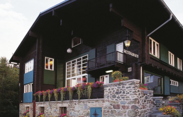 Landhaus Khnuner aus dunklem Holz mit Sockel aus Stein und grünen Fensterläden. Blumen wachsen auf der Terrasse über das Geländer hinab.