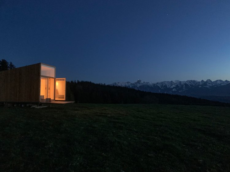 Kleines Tiny House mit beleuchteten Fenstern bei Nacht mit Ausblick in die Berge.