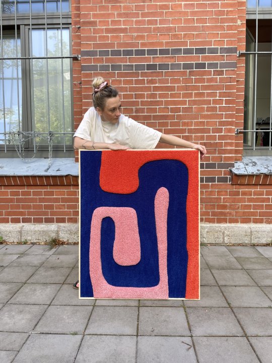 Corinna Müller steht vor einer Backsteinwand und präsentiert einen ihrer Wandteppiche mit geschwungenen Farben in Blau, Rosa und Rot.