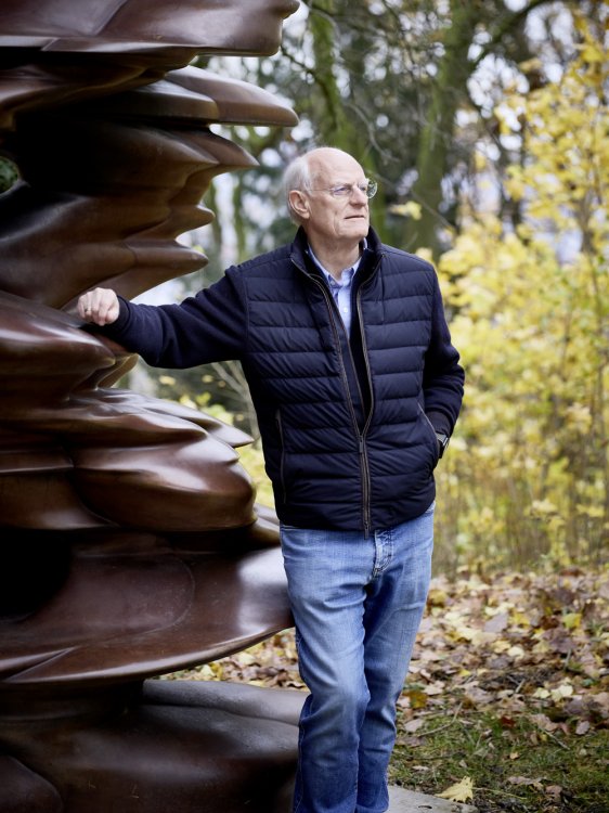 Porträt des britisch-deutsche Künstler Tony Cragg, der sich mit einer Hand auf eine organische Bronzeskulptur stützt, im Hintergrund ein gelber Laubwald.