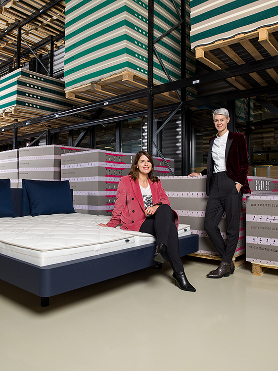Die Schwestern Reta und Iva Schnellmann in der Produktionshalle von Riposa, Reta sitz auf einem Bett, Iva steht daneben.