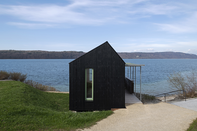 Aussenansicht des Tiny House, ein schwarzer Monolith mit Giebeldach und See im Hintergrund.