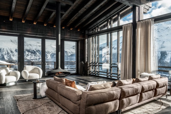 Blick ins Wohnzimmer in einem modernen Chalet mit hängendem schwarzen runden Kamin, braunen Polsterlandschaft und Blick auf eine Winterlandschaft in den Bergen.