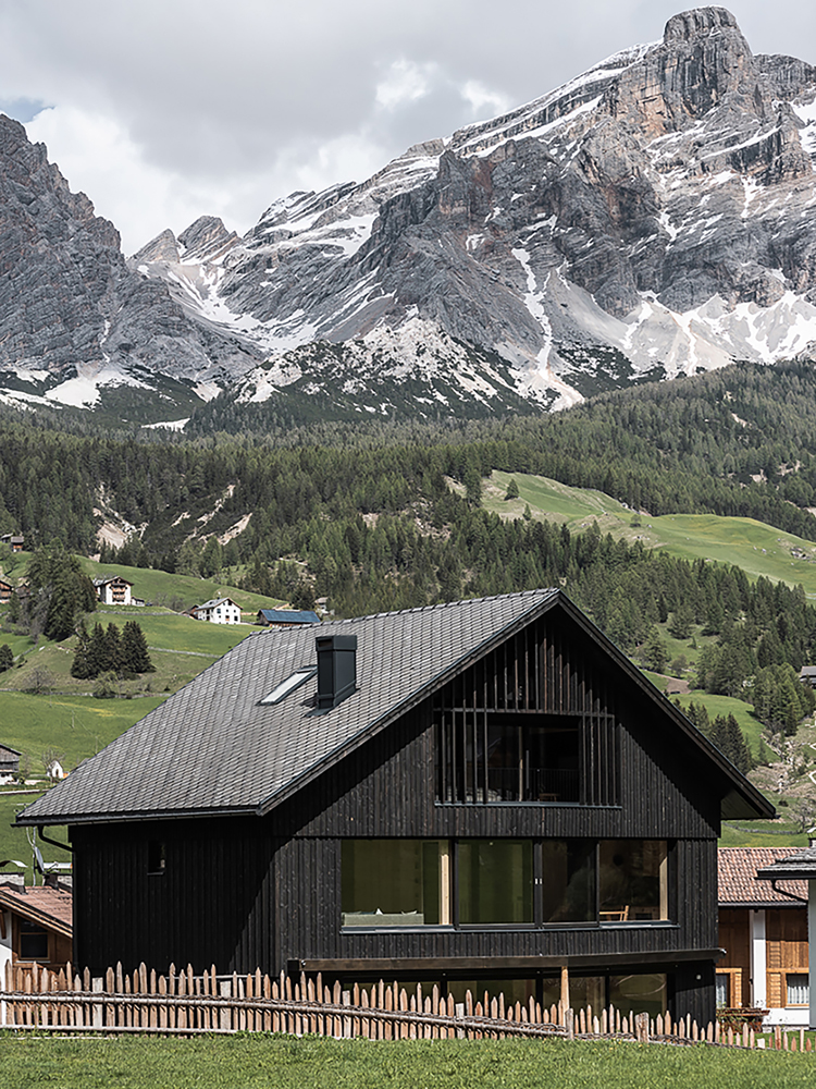 Das dunkle moderne Holzhaus Larix Lodge steht im grünen umrahmt von einem imposanten Bergpanorama der Dolomiten.