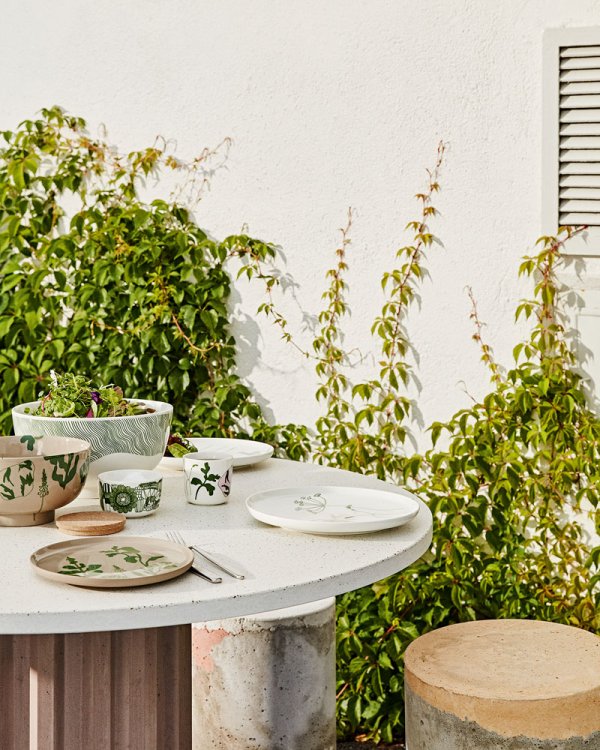 Runder Gartentisch dekoriert mit diversem weissem Geschirr und grünen Pflanzen im Hintergrund.