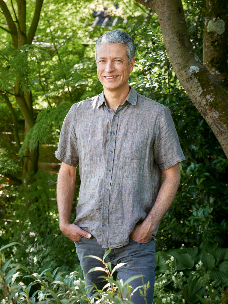 Mann mit grauem Shirt steht inmitten der Natur vor einem Baum.