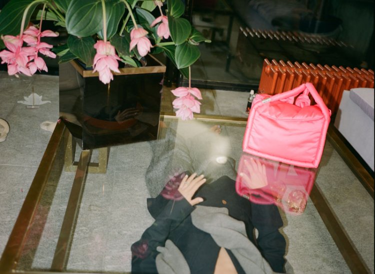 Neon pinke quadratische Handtasche aus weichem Stoffmaterial, die auf einen Beistelltisch aus Glas neben einem rosa Tulpenstrauss steht.