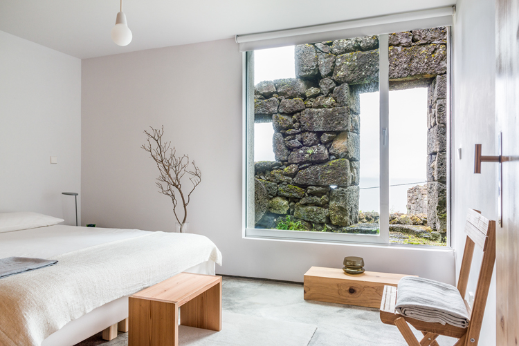 Schlafzimmer mit Bett auf der linken Seite und weissem Bettbezug und einem Panoramafenster, von wo aus man die gegenüberliegende Hauswand mit groben Steinen sieht.