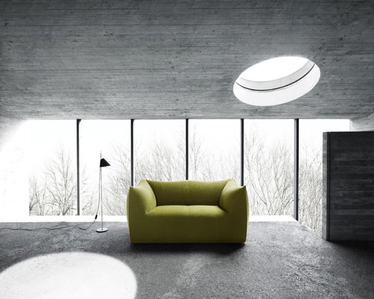 Ein grünes Zweisitzer-Sofa von vorne fotografiert. Steht vor einer Fensterwand in einem Betonraum. Hintergrund in grau-weiss-schwarz-Tönen gehalten, so dass die grüne Farbe heraussticht.