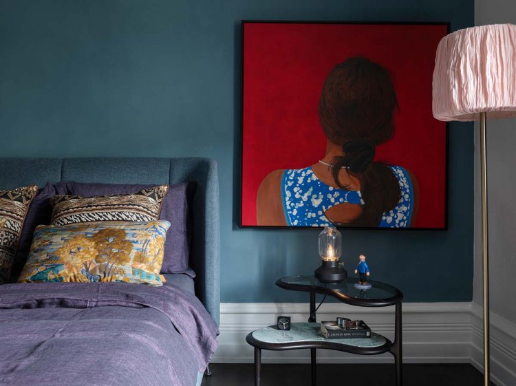 Ein Foto des Schlafzimmers. Die Wand ist blau bemalt und an ihr, direkt neben dem Bett, hängt ein Gemälde einer Frau mit langen schwarzen Haaren.