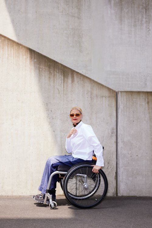 Eine Frau im Rollstuhl welche ein Hemd und eine Jeans trägt mit Sonnenbrille.