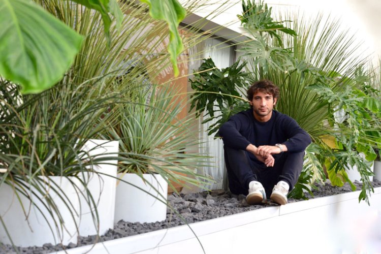 Der Architekt Derek Castiglioni, 35, portraitiert auf einer weissen Mauer sitzend, umgeben von vielen Pflanzentöpfen.