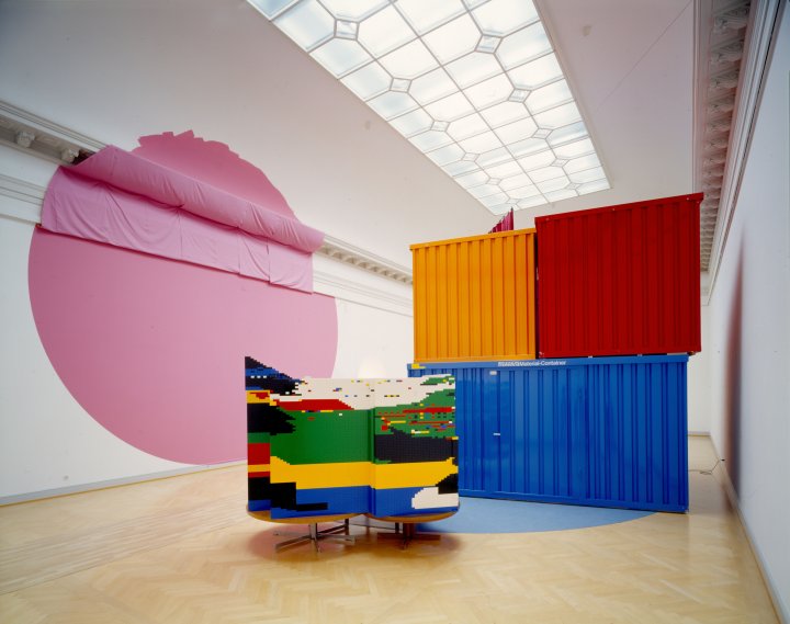 Saalansicht der Ausstellung Citta irreale, links an der Wand ist ein grosser rosa Kreis gemalt, im Raum stehen ein blauer, roter und gelber Container, davor ein buntes Objekt.