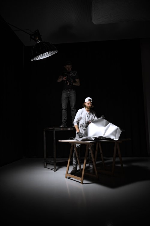 Der Künstler und Möbeldesigner Fabian Bolliger steht in einem dunklen Raum hinter einem Tisch gebeugt und zieht ein Papier von einer Fläche ab, dahinter steht ein Mann mit einer Kamera auf einem Tisch, links ragt eine grosse Lampe ins Bild, die die Situation beleuchtet.