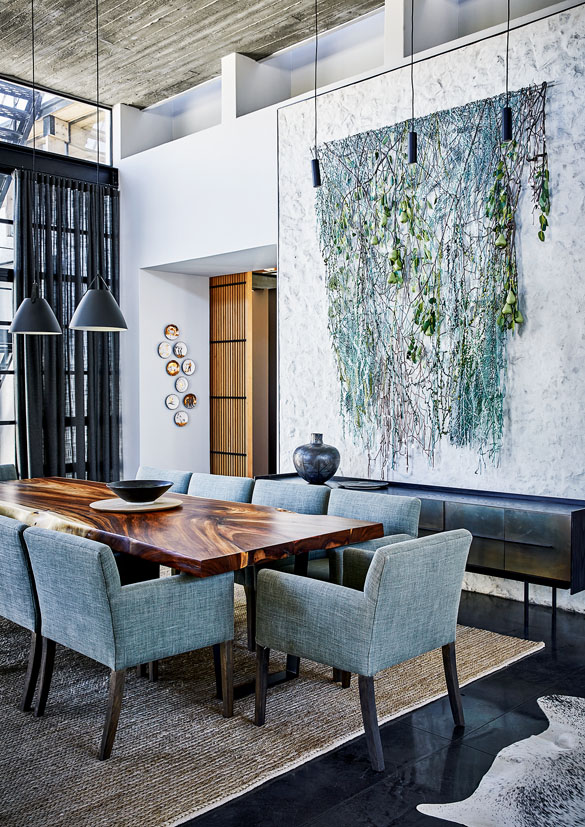 Ein hoher Wohnraum im Industrie-Style mit einem Esstisch aus dunklem Holz, dazu Stühle in grau-bläulichem Stoffbezug, an der Wand hinter dem Tisch hängt ein modernes Kunstwerk aus hinabhängenden Pflanzen.