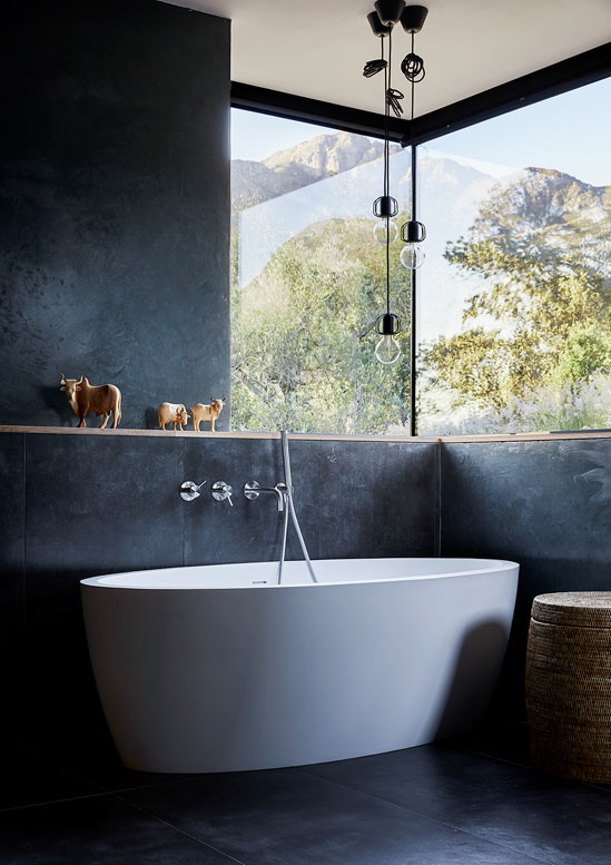 Eine freistehende Badewanne steht im Eck eines modernen, schwarzen Badezimmers, darüber gibt ein grosses Eck-Panoramafenster den Blick auf eine dahinterliegende Berglandschaft frei.