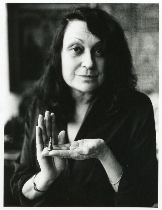 Schwarz-Weiss Porträt der Architektin Lina Bo Barde, die ihre linke Hand zur Kamera streckt, auf der sie ein kleines Objekt präsentiert.