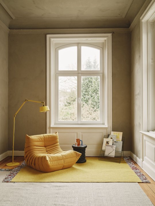 Der gelbe Sessel "Togo" von Ligne Roset auf einem knallgelben Teppich steht in einem ockerfarben gestrichenen Raum in einer Altbauvilla vor einem Fenster, links über den Sessel beugt sich eine gelbe Leselampe.