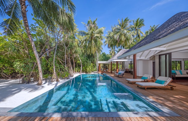 Ein meterlanger Pool entlang einer Holzterrasse mit Sonnenliegen in einer der Family Beach Pool Villas ist umgeben von hohen Kokospalmen und weissem Sand.