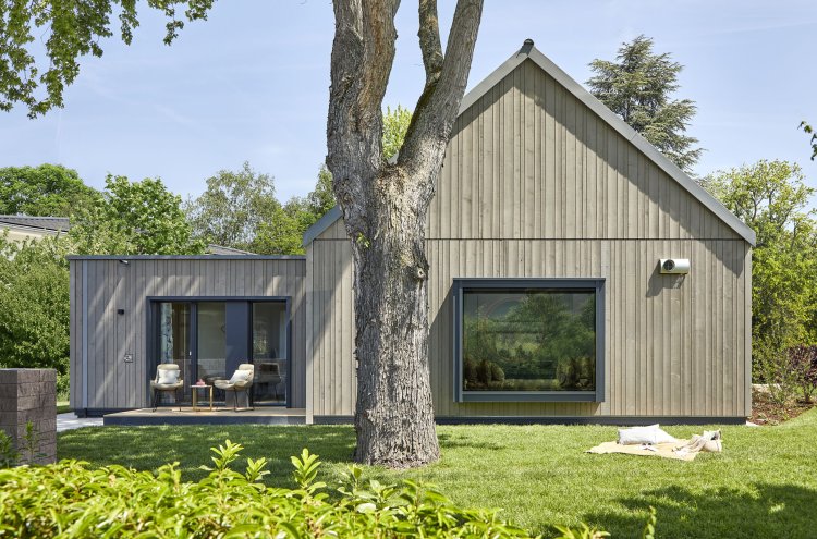 Rückenansicht des neuen Musterhauses Freiraum von Baufritz mit grauer Fichtenfassade und lauschigem Sitzplatz in einem grünen Garten.