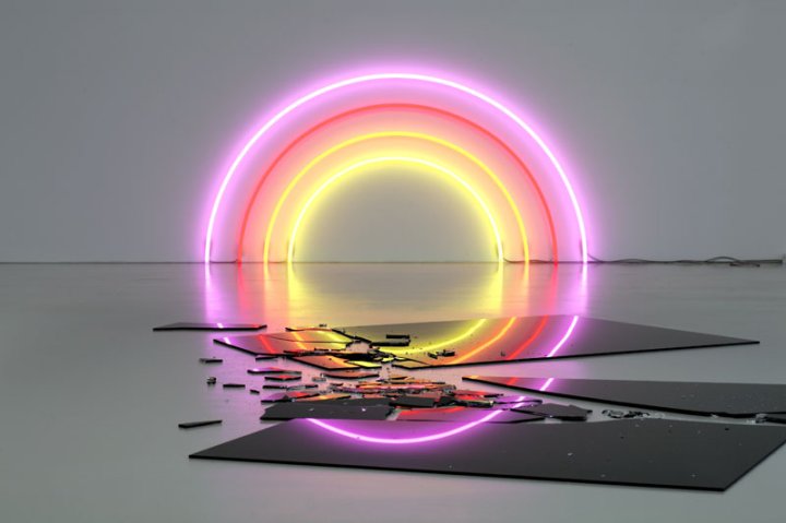 Eine Kunstinstallation aus gebogenen Neonröhren in rosa, rot, orange und gelb die einen Regenbogen bilden von der Künstlerin Lori Hersberger leuchtet in einem Ausstellungsraum, davor liegen zerbrochene, schwarz glänzende Platten.