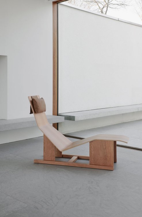 Eine moderne Chaiselongue komplett aus Holz geformt in einem modernen Betonwohnraum vor einer grossen Fensterfront.