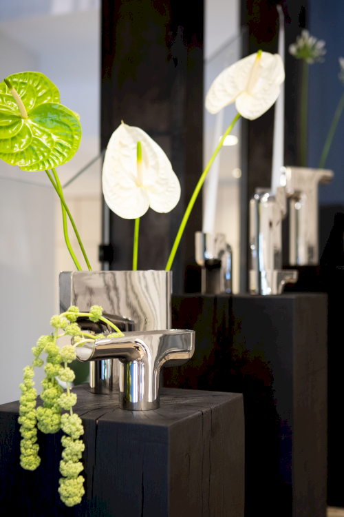 Verschiedene Silbervasen mit exotischen Blumen stehen hintereinander aufgereiht auf schwarzen Holzsockeln.