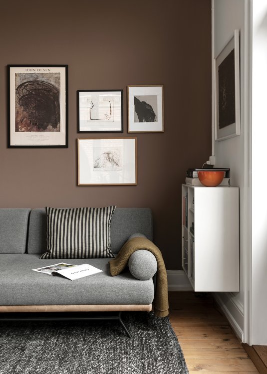 Ein graues Daybed auf dem ein aufgeschlagenes Buch und eine braune Decke liegen steht vor einer braun gestrichenen Wohnwand, an der 4 unterschiedlich grosse abstrakte Bilder und Zeichnungen hängen.
