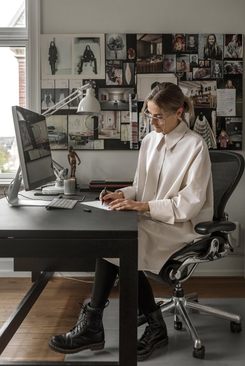 Line Nevers Krabbenhøft, Head of Design bei Sofacompany, sitzt in einer weissen Bluse und mit Brille an einem schwarzen Schreibtisch und skizziert, hinter ihr ist ein Moodboard mit diversen Bildern und Fotos zu sehen.