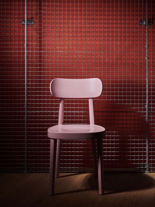 Ein rosaroter Thonet-Stuhl steht vor einer rotbemalten Wand mit einem Gitter davor und ist spärlich beleuchtet.