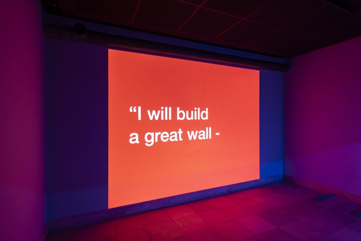 Eine rot hinterleuchtete Wand des Künstlers Tony Cokes auf der die Worte "I will build a great wall" in weisser Schrift stehen, leuchtet in einem Ausstellungsraum.
