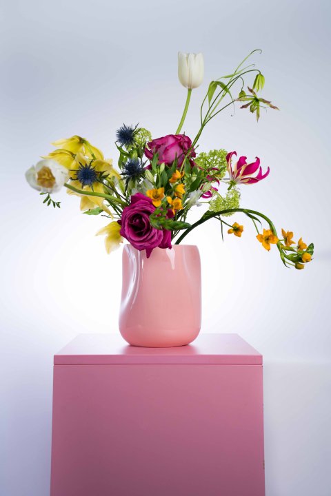 Eine rosafarbene Vase mit einem frühlingshaften Blumenbouquet steht auf einem rosafarbenen Sockel.