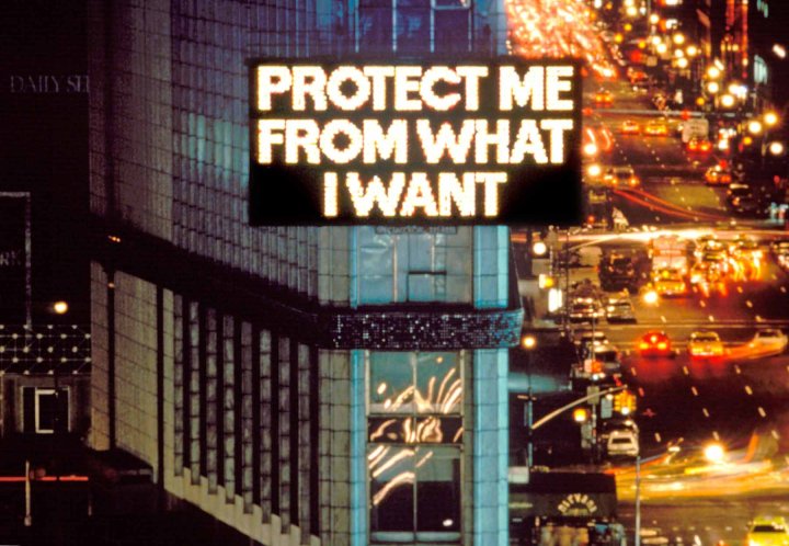 Ein LED-Kunstwerk der Künstlerin Jenny Holzer mit dem Schriftzug "Protect me from what I want" in Grossbuchstaben hängt an einem Hochhaus in einer Stadt bei Nacht.