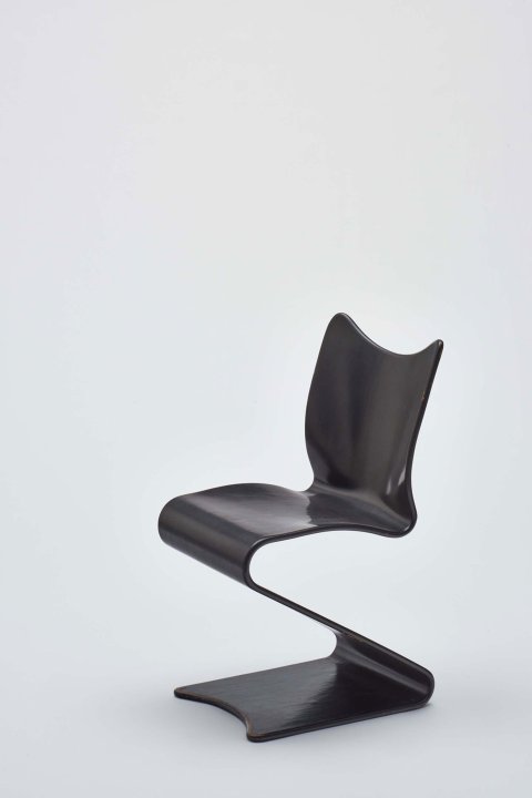 Ein aus einer Holzplatte geformter schwarzer Stuhl, dessen Rücken- und Sitzfläche in gefalteter Weise in Beine übergehen.