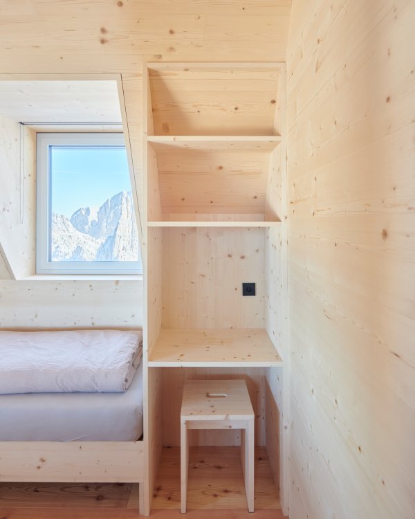 Man sieht eine Holzablage, ein Hocker und ein Bett sowie den Blick aus dem Fenster in die Berge.