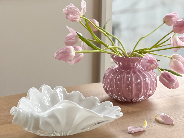 Ein Tisch mit einer weissen Glasschale in Muschelform. Rosarote Tulpen in einer rosa Vase auf einem Holztisch.