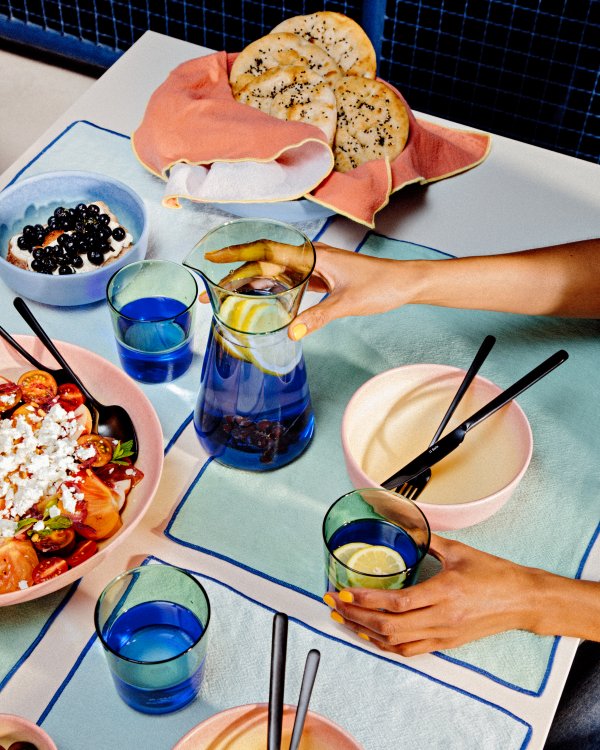 Ein sommerlicher Tisch mit Essen in bunten Tellern und Händen, die zu Gläsern und Besteck greifen.